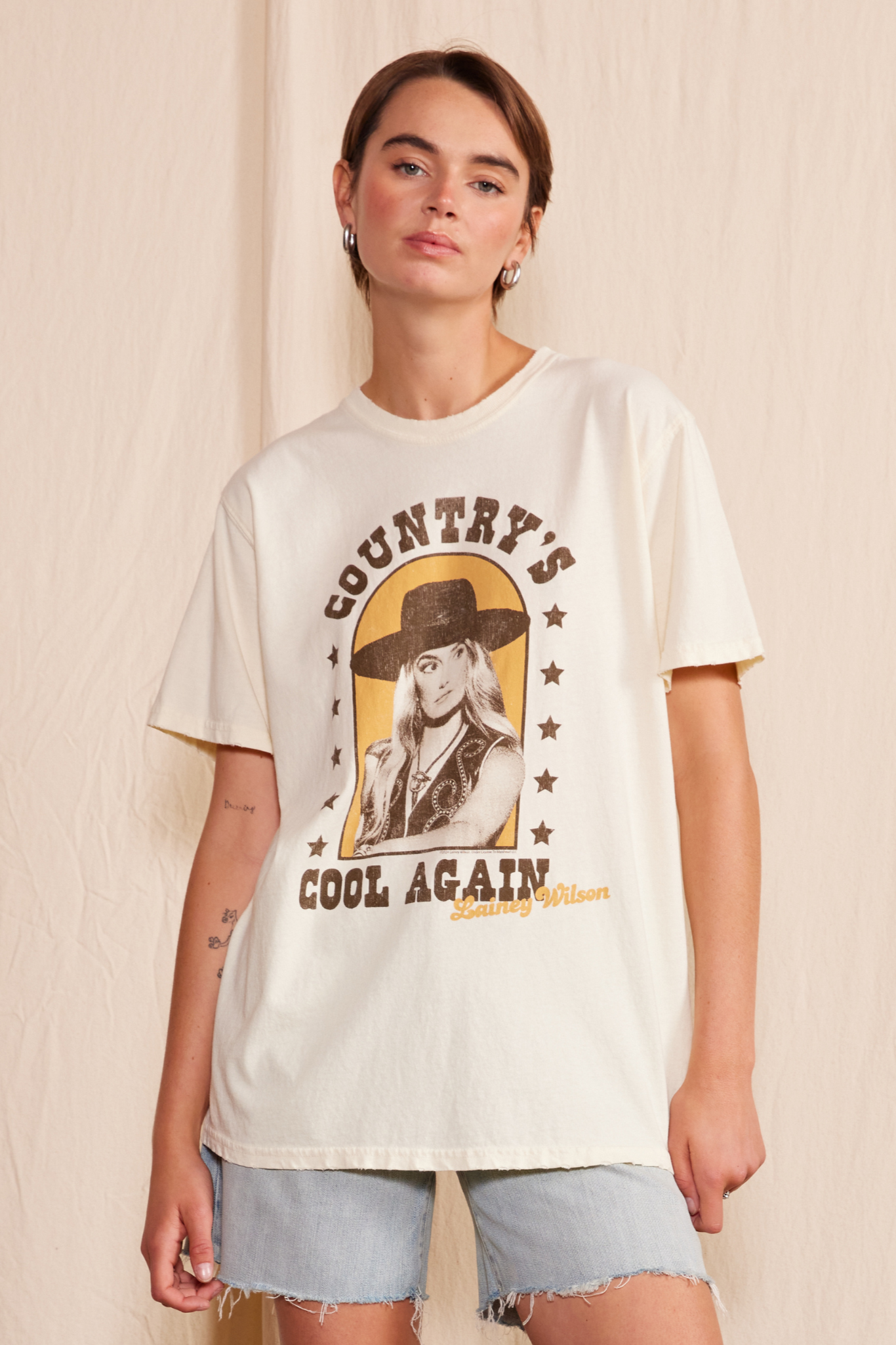 Unisex off-white tee featuring Lainey Wilson with brown stars and text that reads “COUNTRY’S COOL AGAIN.”