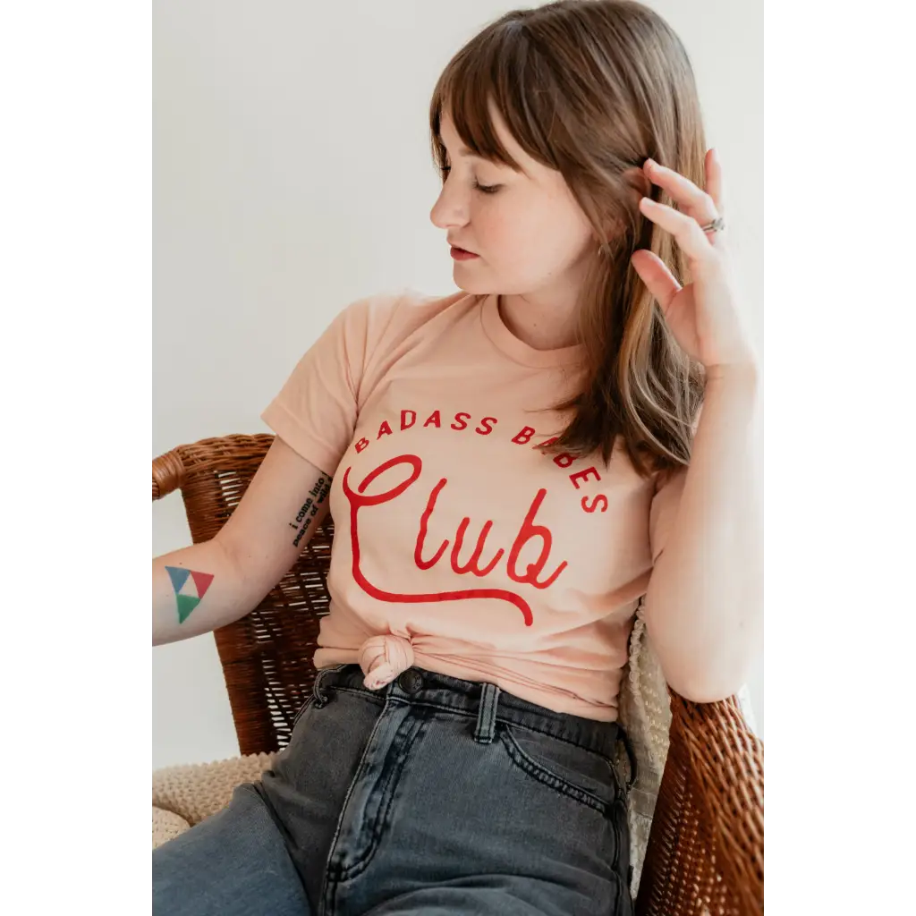Relaxed female model wearing peach t-shirt with red text that reads, “BAD ASS BABES Club.”