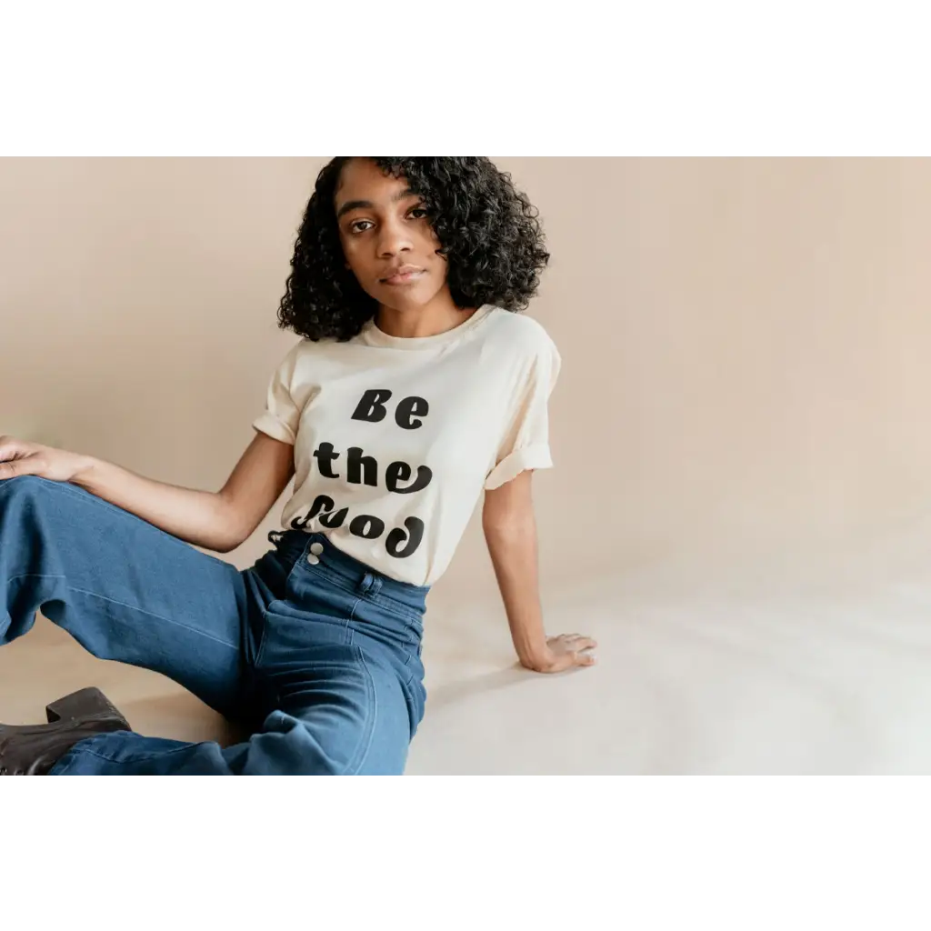 Model seated casually seating ivory t-shirt with “be the good” in black text