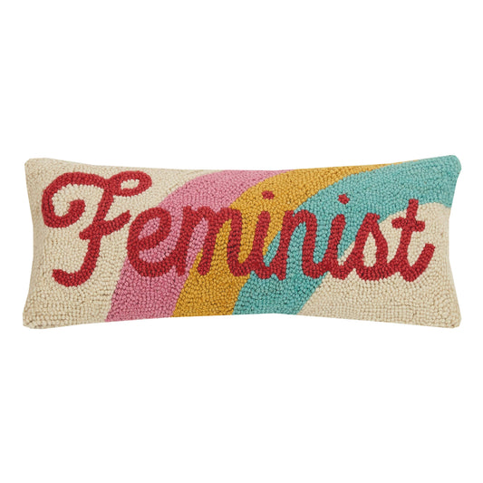 Lumbar shaped accent pillow with rainbow design against red writing “feminist” in cursive letters