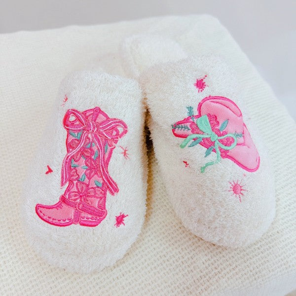 Fuzzy pair of white slippers with pink embroidered western boot and cowgirl hat designs on a white background