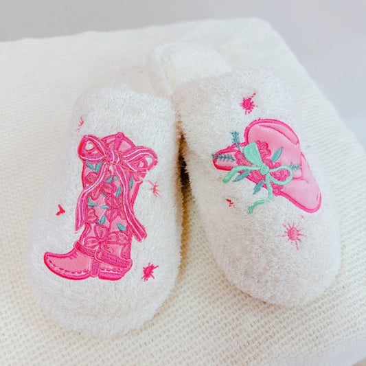 Fuzzy pair of white slippers with pink embroidered western boot and cowgirl hat designs on a white background
