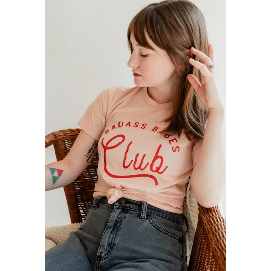 Relaxed female model wearing peach t-shirt with red text that reads, “BAD ASS BABES Club.”