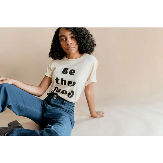 Model seated casually seating ivory t-shirt with “be the good” in black text