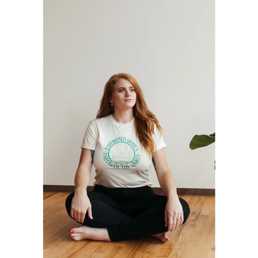 Model seated wearing a white t-shirt with teal text that form a circle that reads “ empowered women empower the world.” 