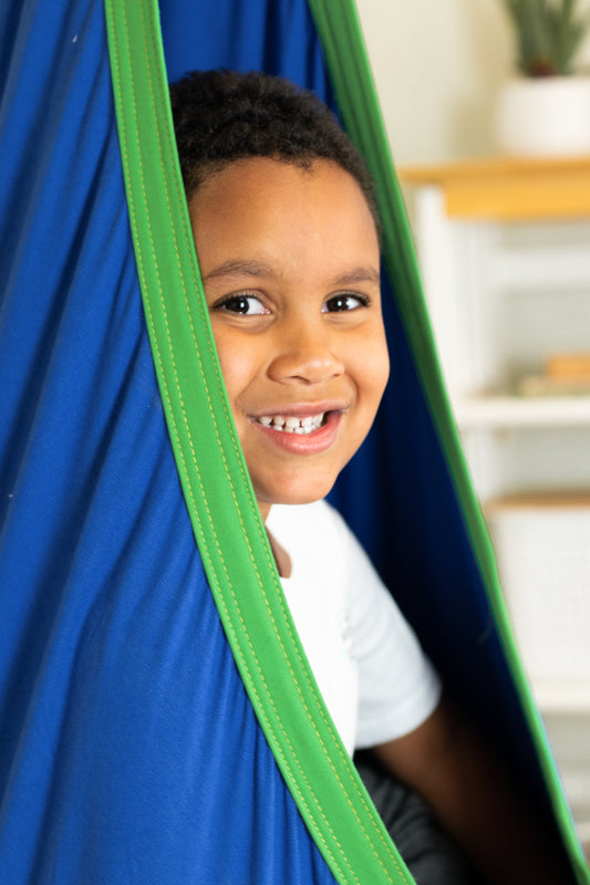 Young boy smiling while seated in Harkla Pod Sensory Swing in blue with bright green trim.