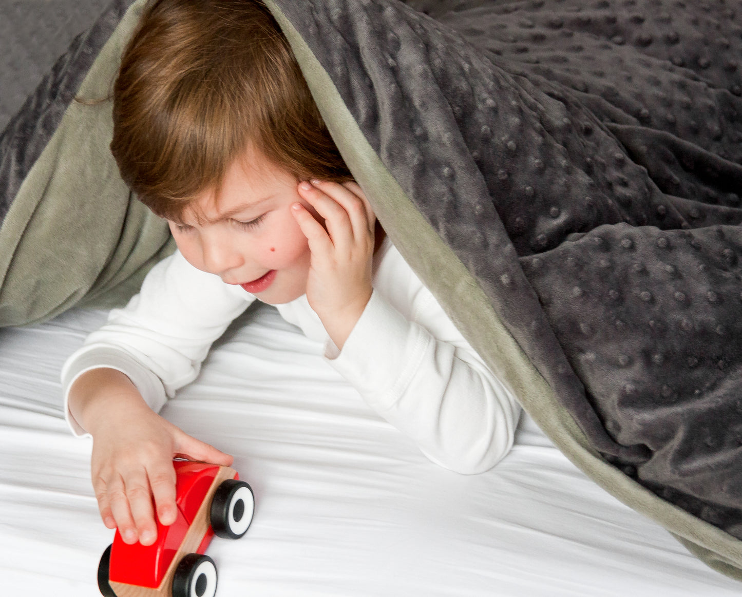 Young boy playing with toy car under calming weighted blanket with velvety charcoal front and soothing gray reverse.