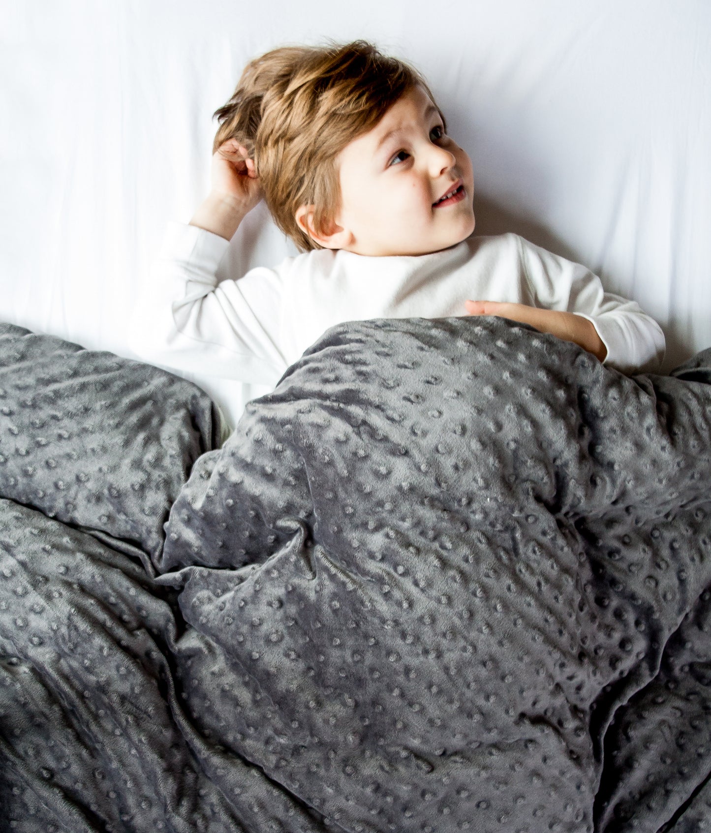 Young boy relaxing under snug Lottie Dottie Weighted Blanket in gray.