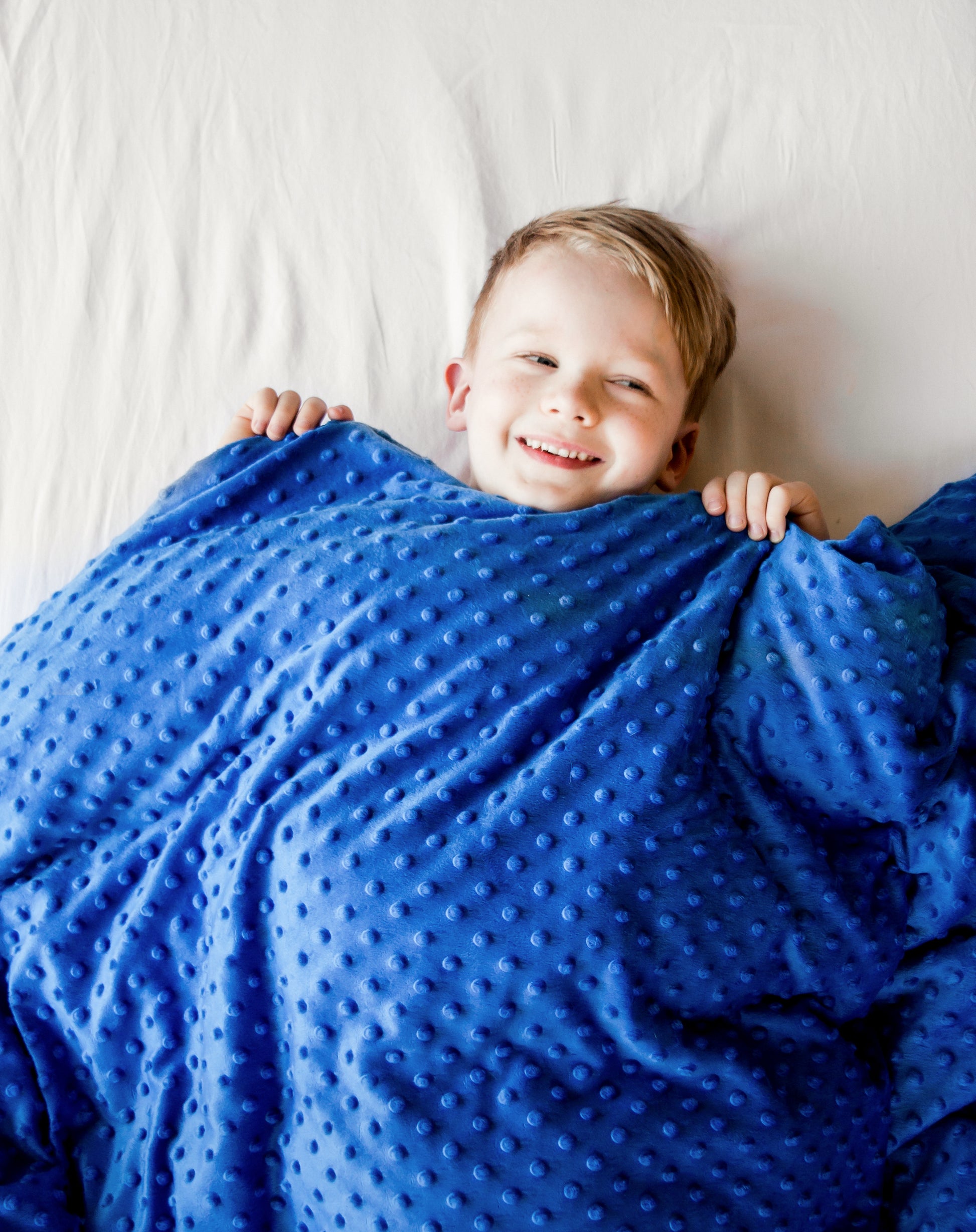 Young boy smiling under cozy, Lottie Dottie Weighted Blanket in bright blue.