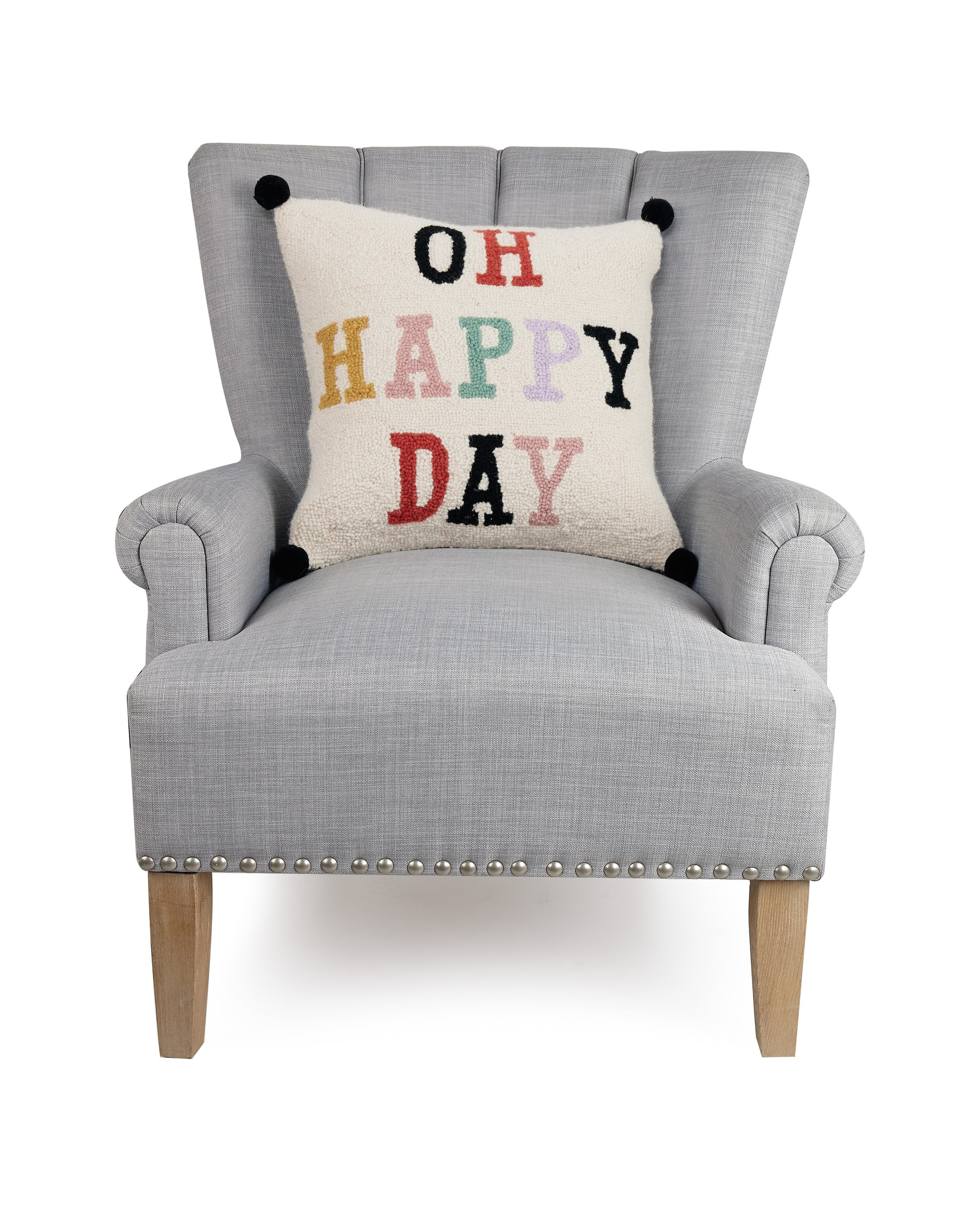 Cream Yvonne Ellen throw pillow with black tassels and multi-colored letters that read, “Oh Happy Day” against a gray upholstered chair.