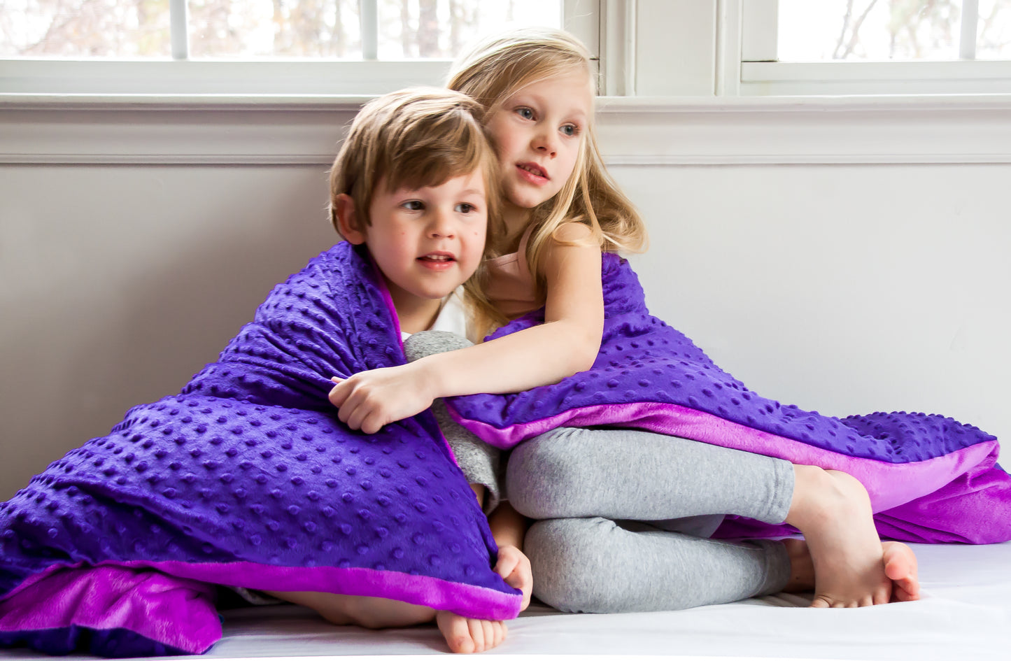 Children snuggling under soothing weighted blanket with purple dotted front and mink lilac reverse.